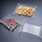 Three decorative glass trays are displayed on a dark surface. One tray holds an assortment of colorful beads, another contains cookies, and the third is empty.